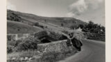 Two men and a child leaning against a bridge. Surrounded my fields in the background.